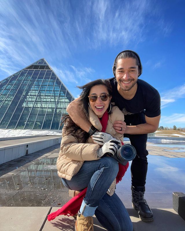 Wil Dasovich in a black t-shirt and pants posing with his sister in a nude jacket and blue jeans holding a camera.
